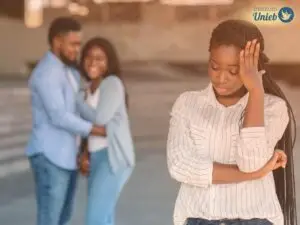Mulher com mão no rosto e outro braço na cintura e ao fundo um casal sorrindo