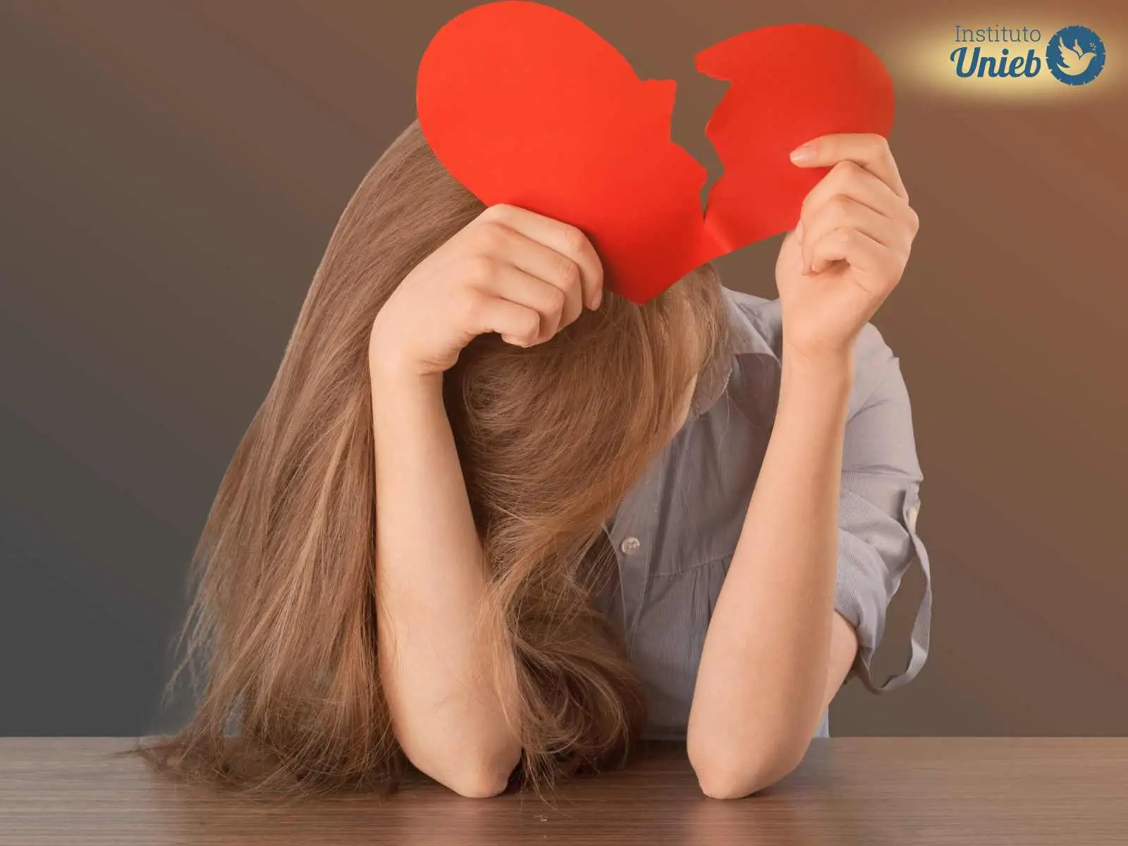 Mulher com a cabeça abaixada e cotovelos apoiados na mesa segurando um coração vermelho partido ao meio