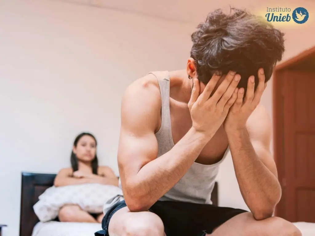 Homem sentado ao pé da cama com as mãos no rosto e cotovelos nos joelhos e mulher ao fundo sentada na cama com expressão de frustração