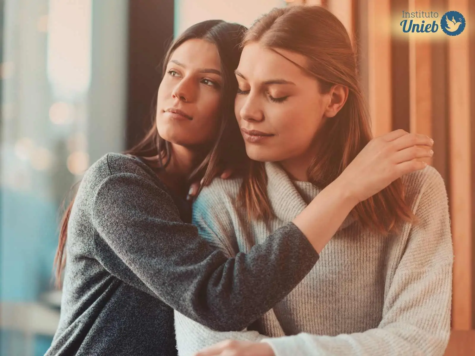 Duas mulheres vestindo roupas neutras abraçadas com expressão pensativa e reflexiva