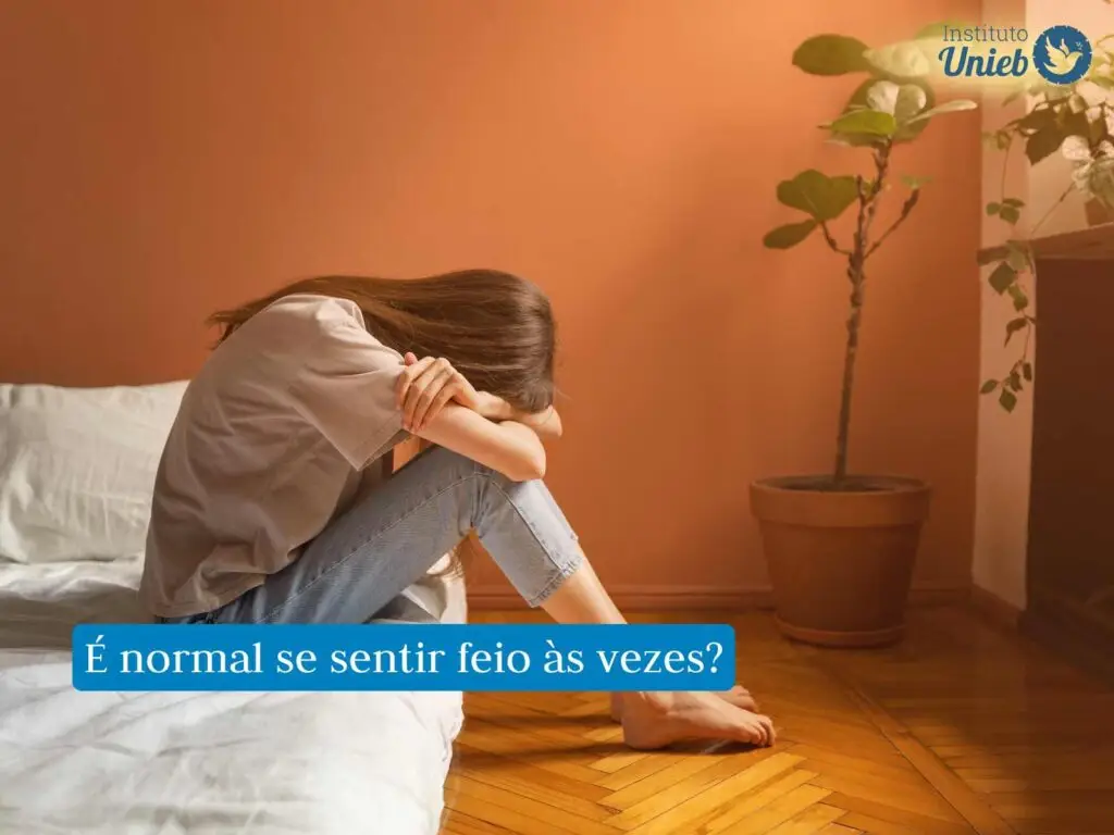 Mulher sentada na cama próxima ao chão debruçada em seus braços e pernas com a cabeça baixa