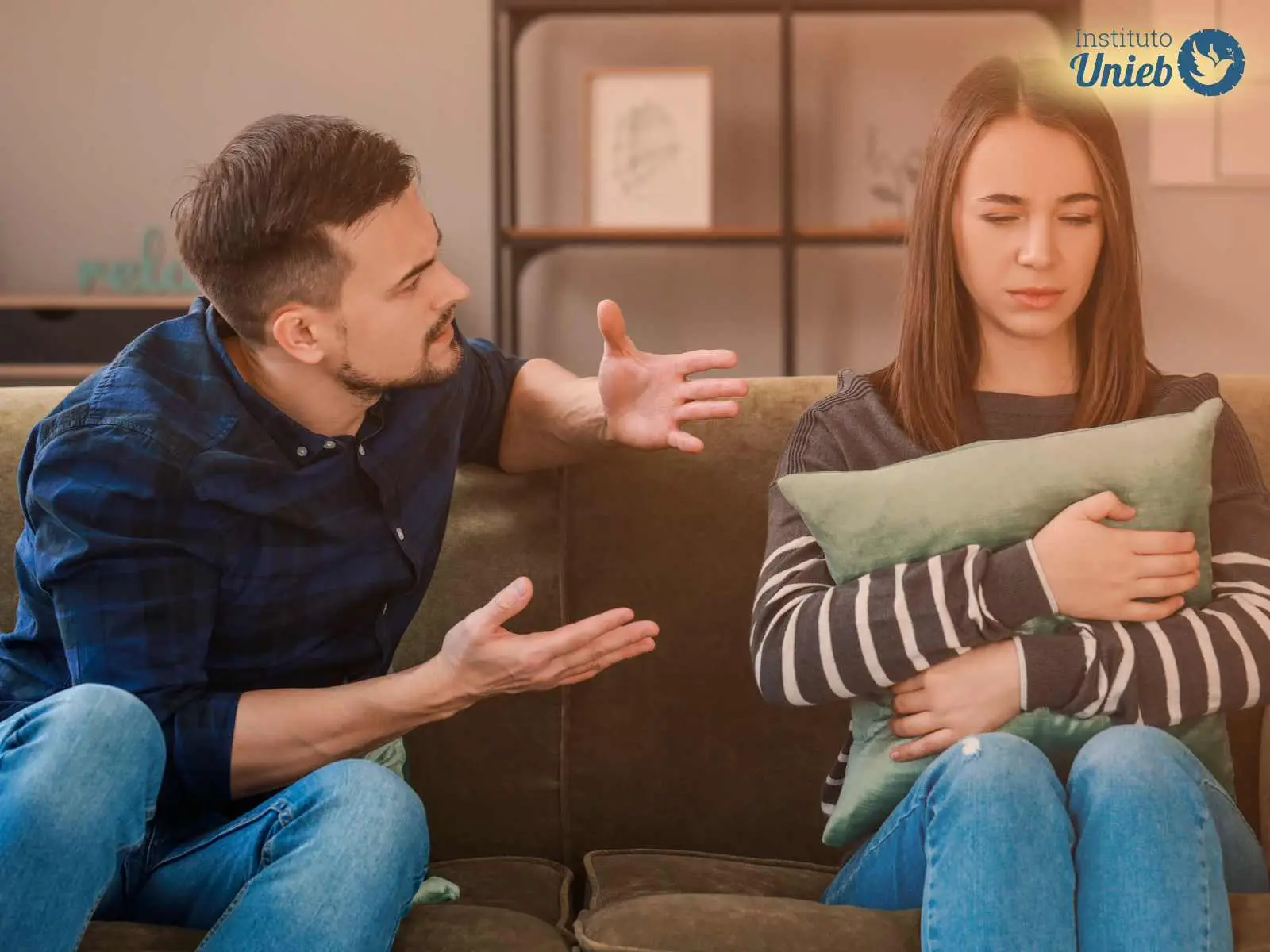 Casal de homem e mulher sentado no sofá, onde mulher está segurando uma almofada próxima ao corpo e homem está gesticulando enquanto briga com sua parceira
