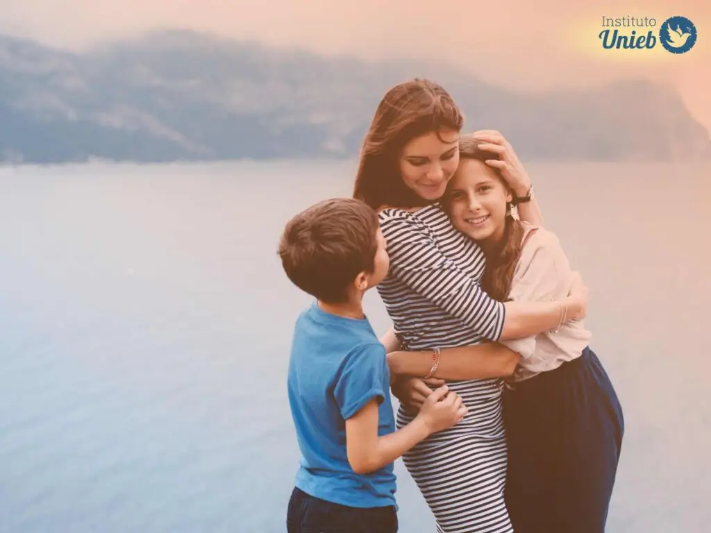 Mulher de vestido listrado abraçando sua filha com filho ao seu lado em momento de carinho