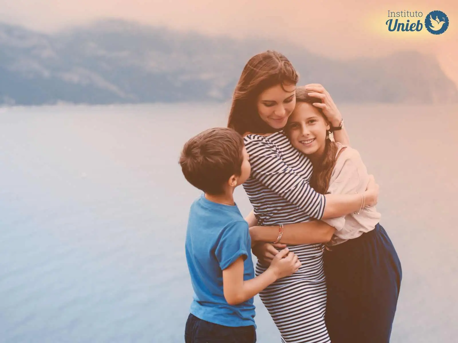 Mulher de vestido listrado abraçando sua filha com filho ao seu lado em momento de carinho