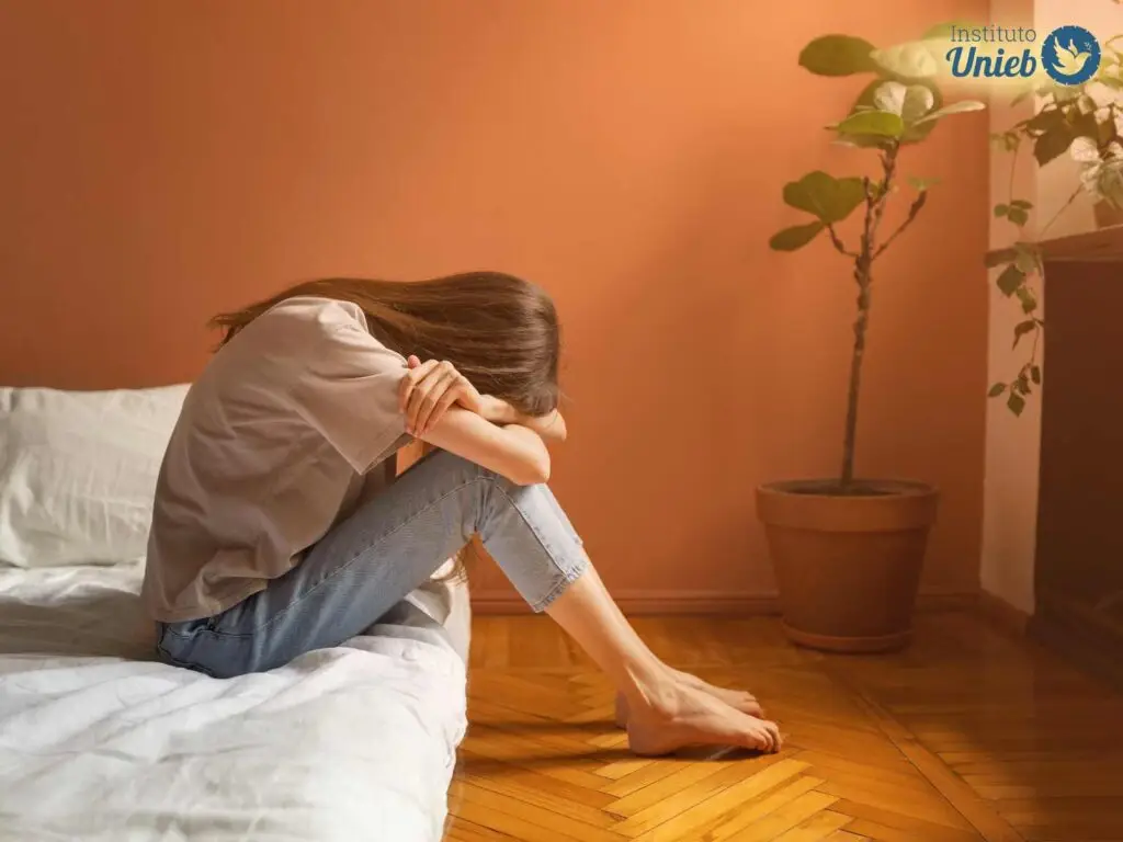 Mulher sentada na cama próxima ao chão debruçada em seus braços e pernas com a cabeça baixa