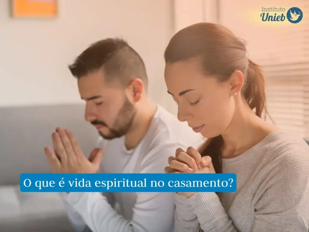 Qual a importância da vida espiritual no casamento? Casal de homem e mulher na sala vestindo roupas em tons de cinza com mãos em prece e olhos fechados rezando