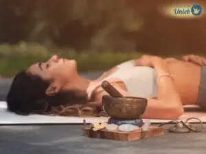 Mulher com top branco deitada no chão com braço em cima do peite e olhos fechados com defumador próximo à câmera e outros elementos de meditação