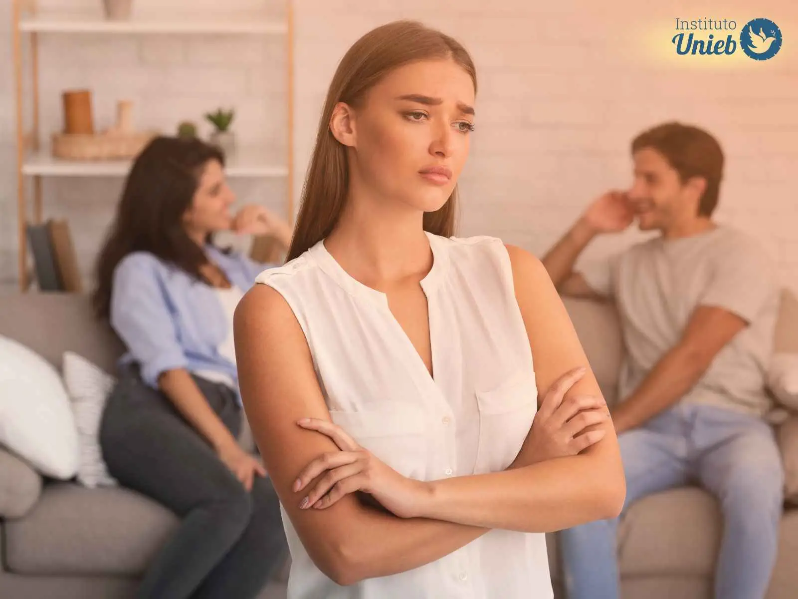 Homem e mulher conversam ao fundo sentados em um sofá e uma mulher vestindo blusa branca com braços cruzados está à frente com expressão de tristeza