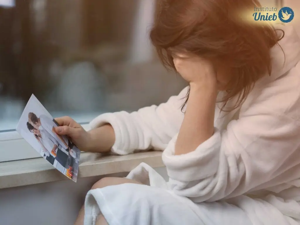 Mulher vestindo roupão branco sentada próximo à janela com fotografia de casal na mão e outra secando lágrimas no rosto