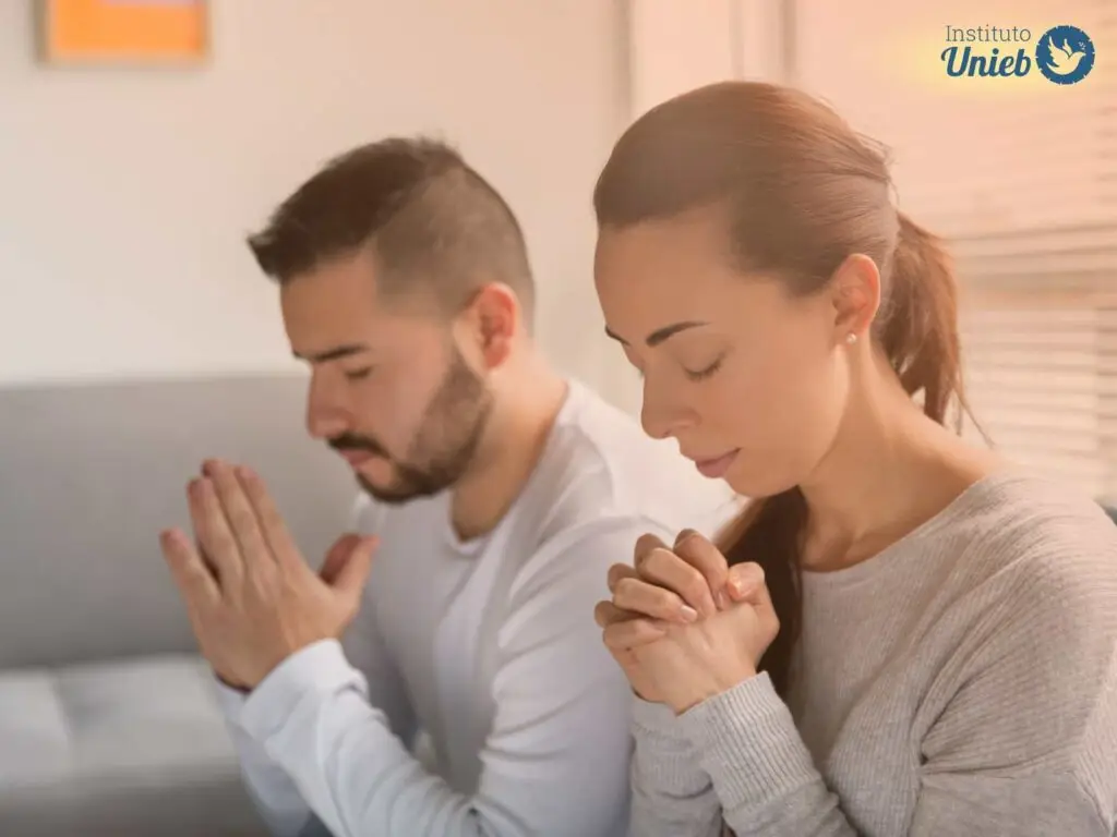 Casal de homem e mulher na sala vestindo roupas em tons de cinza com mãos em prece e olhos fechados rezando