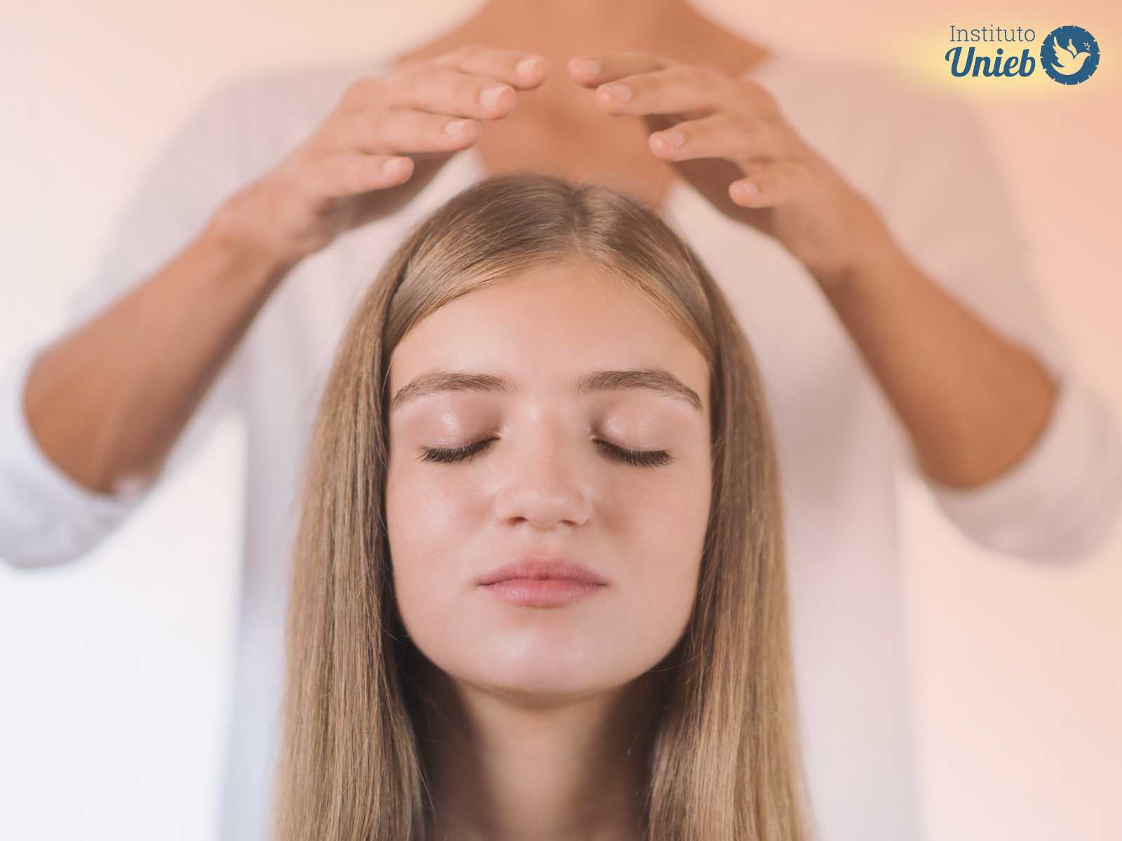 Mulher de cabelo claro e liso sentada com olhos fechados enquanto outra mulher ao fundo coloca as mãos acima da sua cabeça