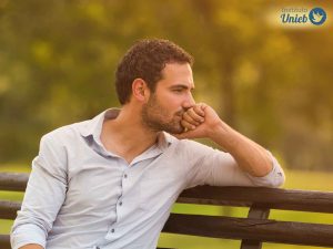 Homem vestindo camisa de botões clara sentado em um banco de madeira ao ar livre com cabeça apoiada em sua mão esquerda enquanto observa pensativo
