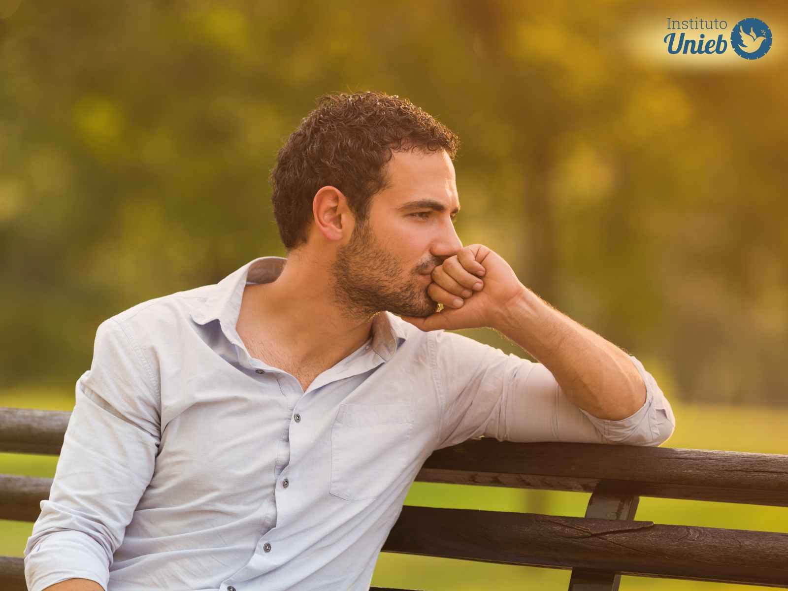 Homem vestindo camisa de botões clara sentado em um banco de madeira ao ar livre com cabeça apoiada em sua mão esquerda enquanto observa pensativo