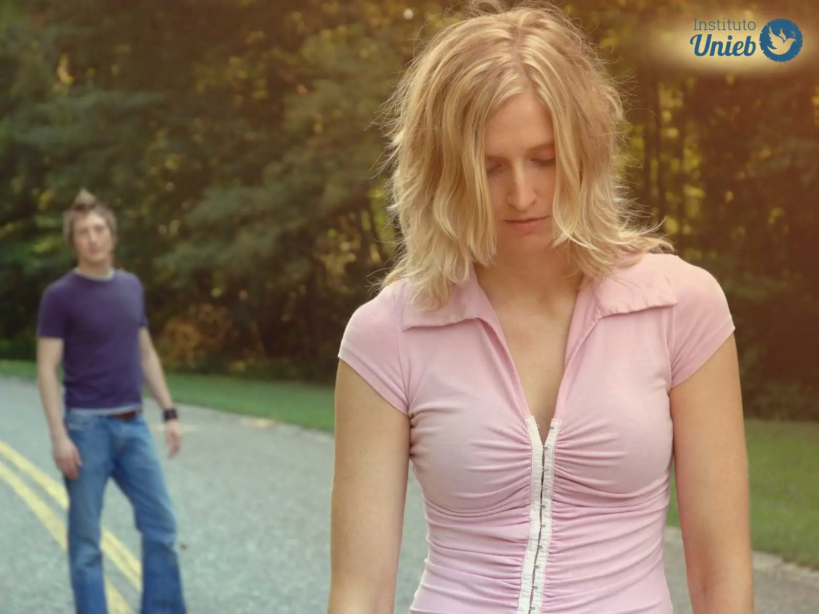 Mulher loira com camiseta rosa mais próxima a câmera com cabeça baixa e expressão triste após ser rejeitada por homem ao fundo que veste camiseta azul e calça jeans