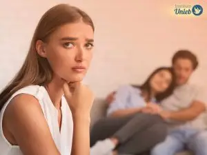 Mulher vestindo regata branca olhando para a câmera com expressão de tristeza e ao fundo um casal se abraça sentado em um sofá