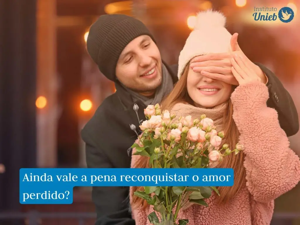 Mulher vestindo sueter e touca de tricô recebendo buquê de flores com os olhos fechados e vedados pela mão de um homem que está atrás  vestindo casado e touca preta