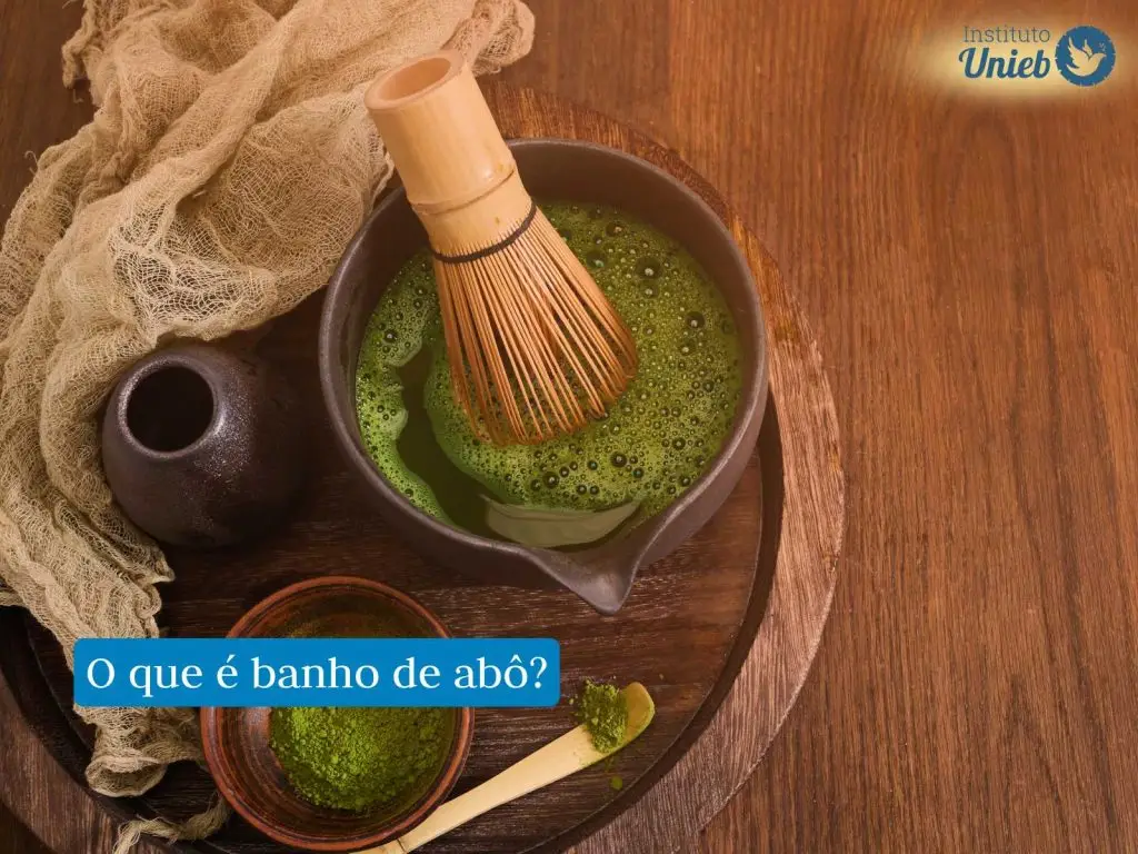 Materiais para a preparação de banho de ervas como um pote com ervas verdes maceradas, outro pote com água com ervas, um tecido próximo aos potes dentro de uma bandeja de madeira em uma mesa de madeira