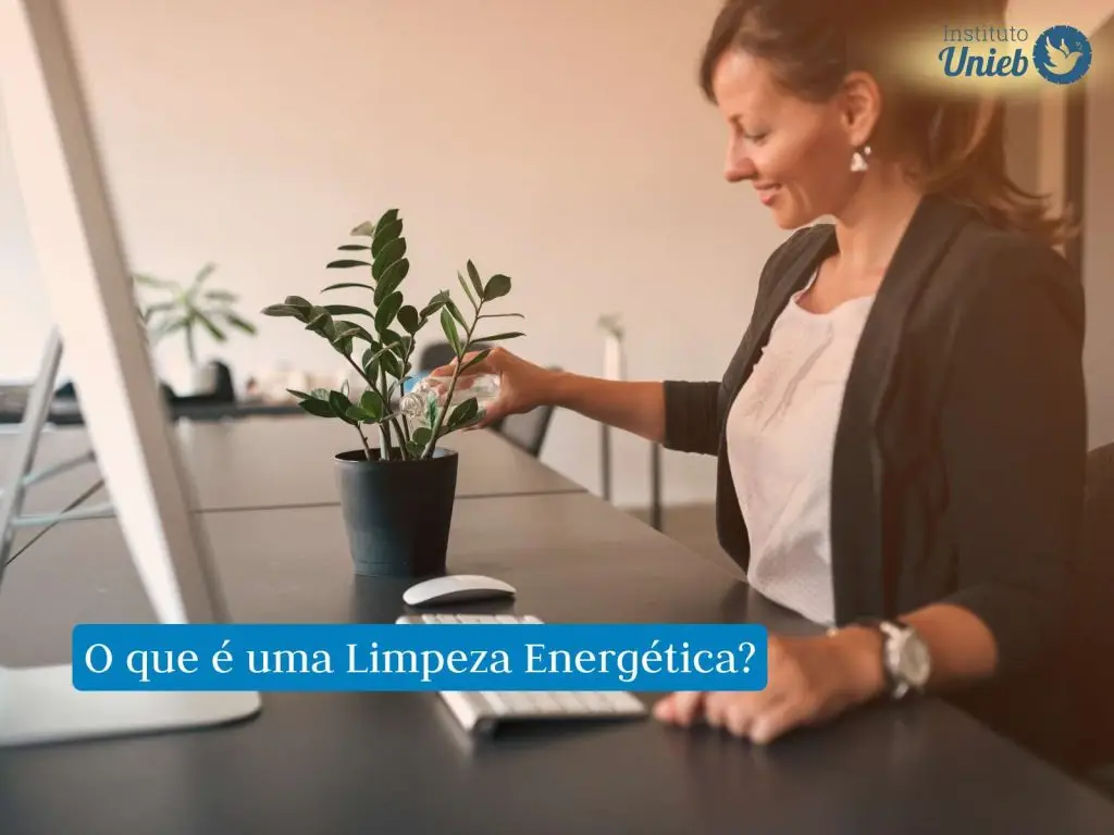 Mulher com roupa formal em local de trabalho regando um vaso de planta sob a mesa de trabalho onde também tem um monitor, um teclado e um mouse
