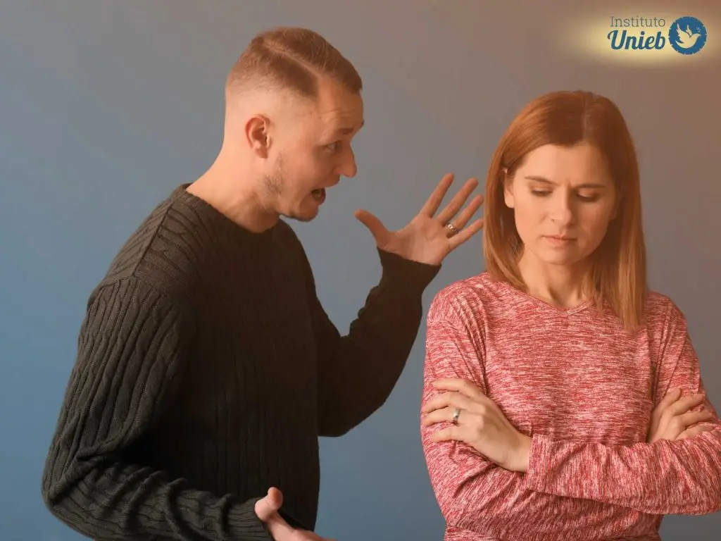 Mulher vestindo blusa rosa olhando para baixo com expressão triste enquanto homem vestindo blusa verde escuro gesticula com a mão esquerda olhando para ela com voracidade