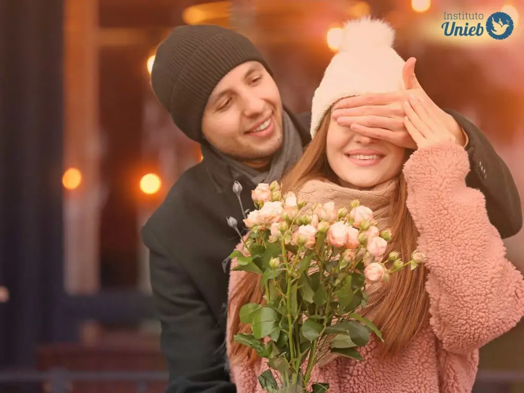 Mulher vestindo sueter e touca de tricô recebendo buquê de flores com os olhos fechados e vedados pela mão de um homem que está atrás vestindo casado e touca preta - Instituto Unieb (Pai de santo Roberson Dariel) - Contato 11 96982-1121 - CEP 14801-040 - Av. Dom Pedro II 562, Centro, Araraquara, SP