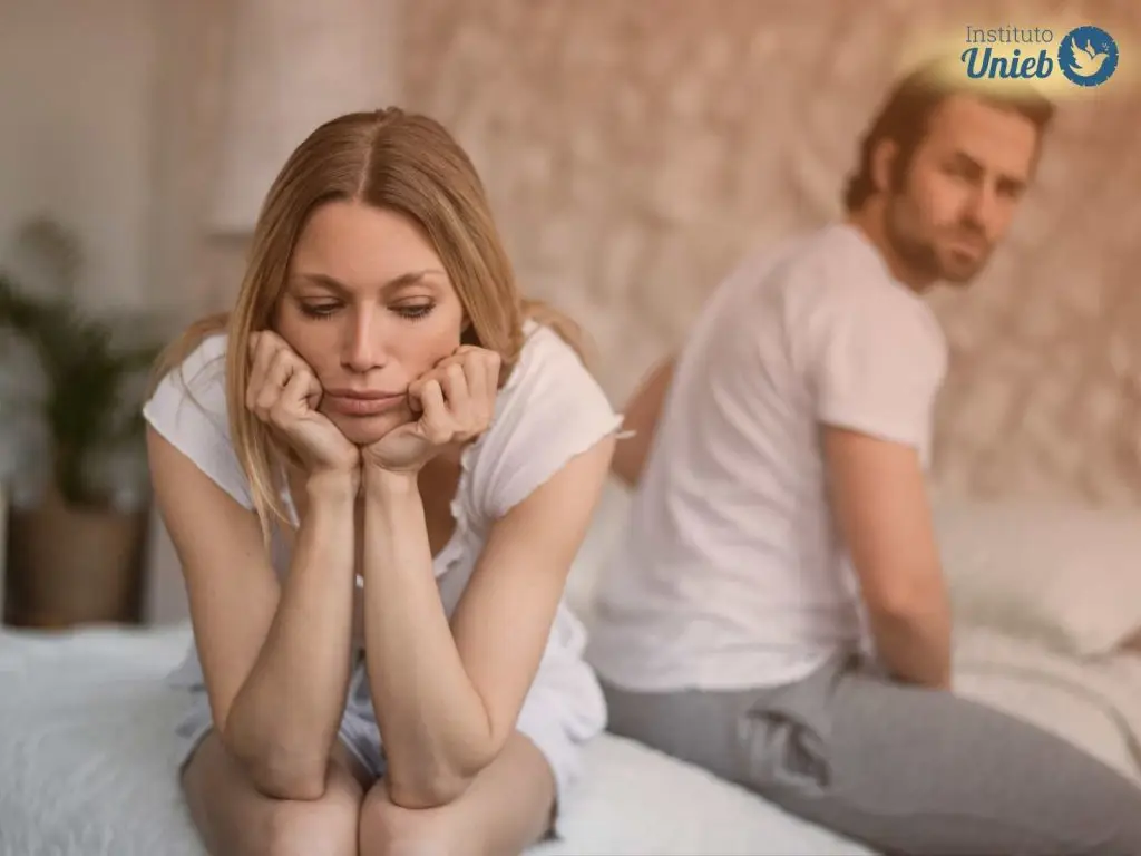 Homem e mulher sentados na cama vestindo camiseta branca com expressão de tristeza