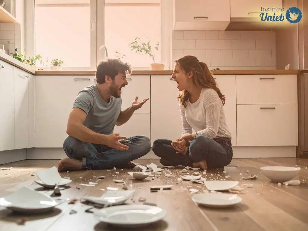 Homem e mulher sentados no chão com pernas cruzadas no meio da cozinha com pratos quebrados no chão em meio a uma discussão