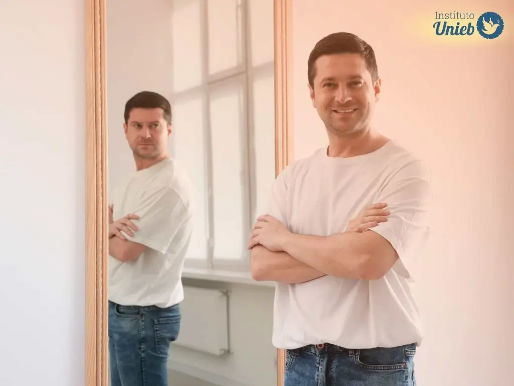Homem vestindo camiseta branca e jeans com braços cruzados olhando para câmera sorrindo com espelho ao fundo refletindo sua imagem com expressão de raiva