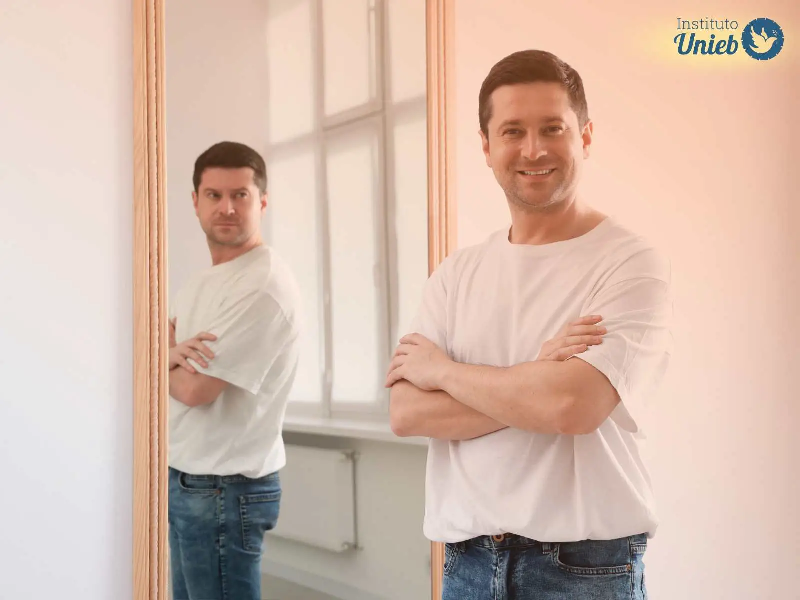 Homem vestindo camiseta branca e jeans com braços cruzados olhando para câmera sorrindo com espelho ao fundo refletindo sua imagem com expressão de raiva