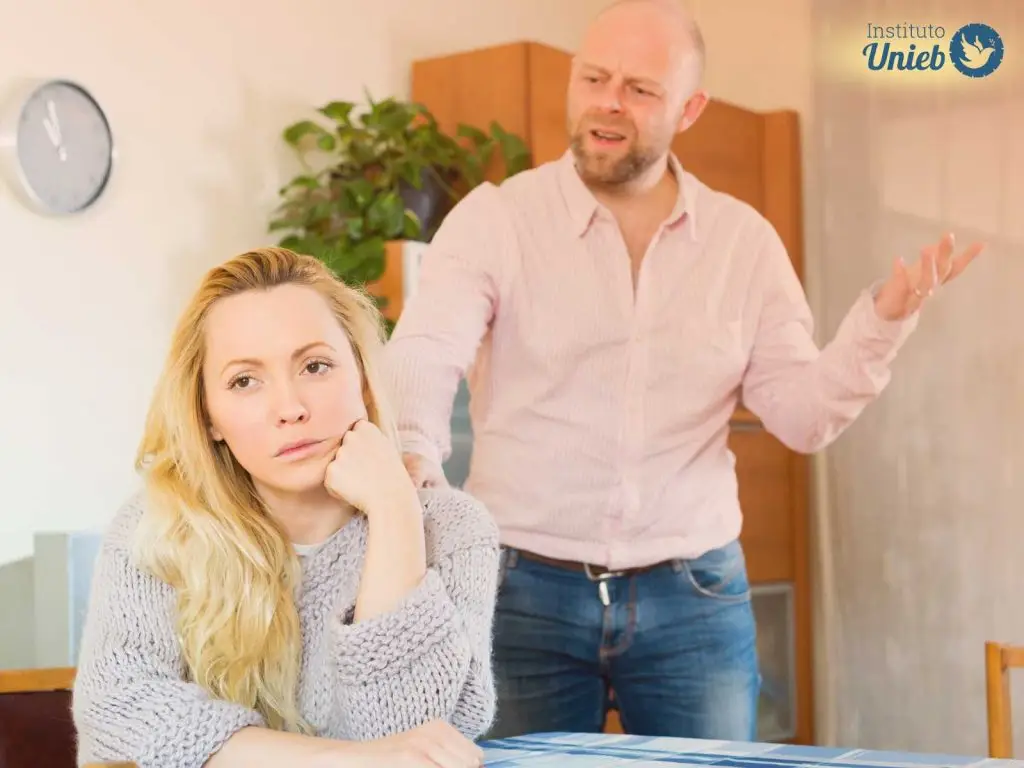 Homem gesticulando irritado e de mau humor atrás da mulher que está sentada com rosto apoiado na mão