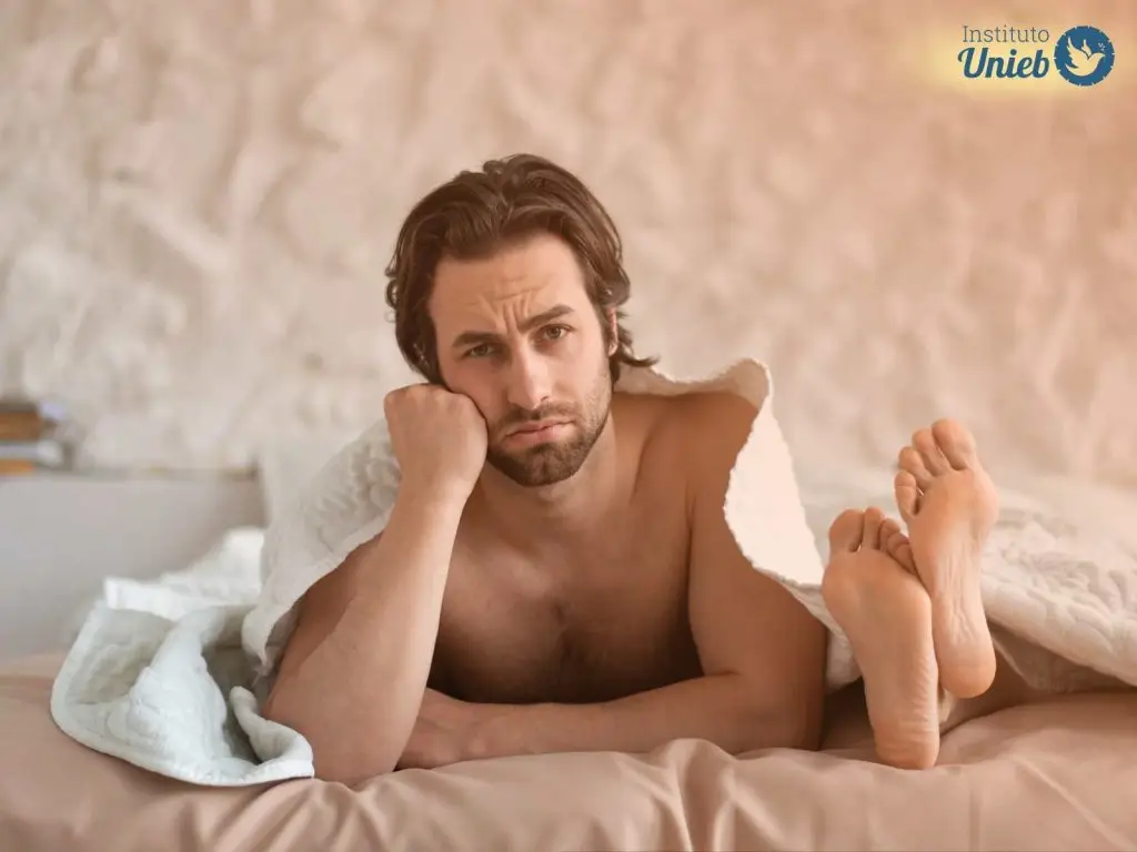 Homem embaixo da coberta deitado na cama com braço apoiado na cabeça e pés da parceira ao lado fora do cobertor