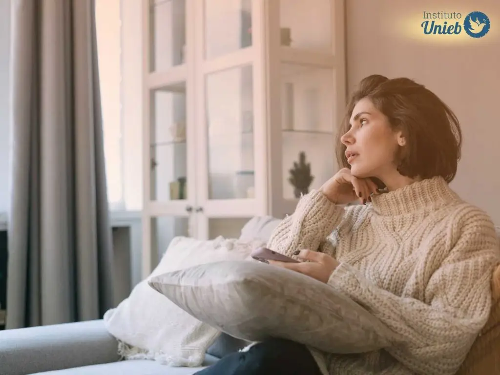 Mulher vestindo sueter de lã bege sentada em sofá da sala com celular em uma mão e a outra com a cabeça apoiada olhando no horizonte