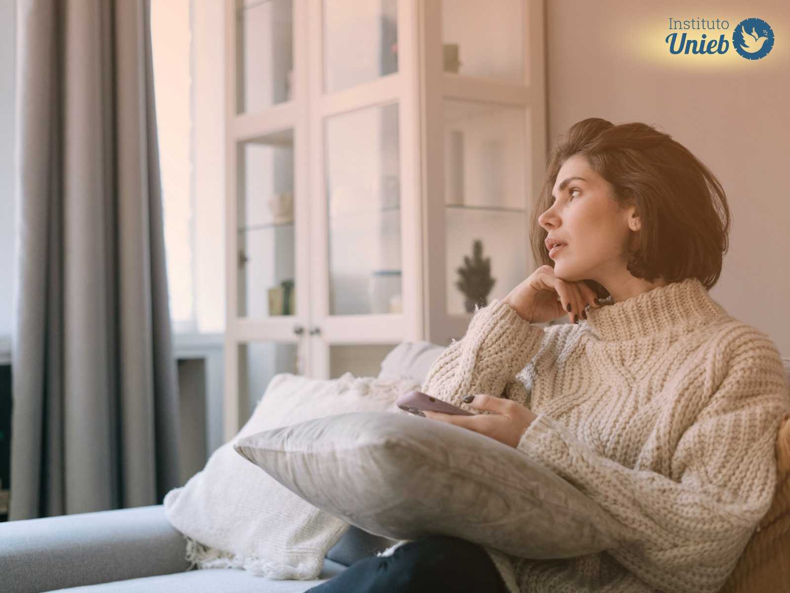 Mulher vestindo sueter de lã bege sentada em sofá da sala com celular em uma mão e a outra com a cabeça apoiada olhando no horizonte