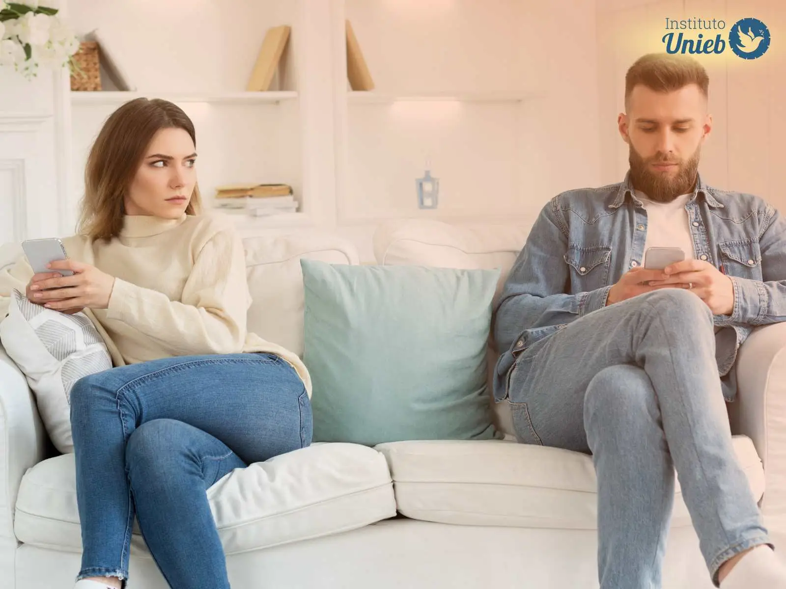 Homem e mulher sentados no sofá distantes um do outro cada um com um celular, a esposa olha para o marido com desconfiança