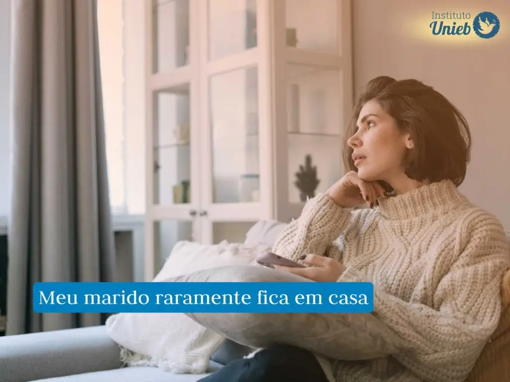 Mulher vestindo sueter de lã bege sentada em sofá da sala com celular em uma mão e a outra com a cabeça apoiada olhando no horizonte