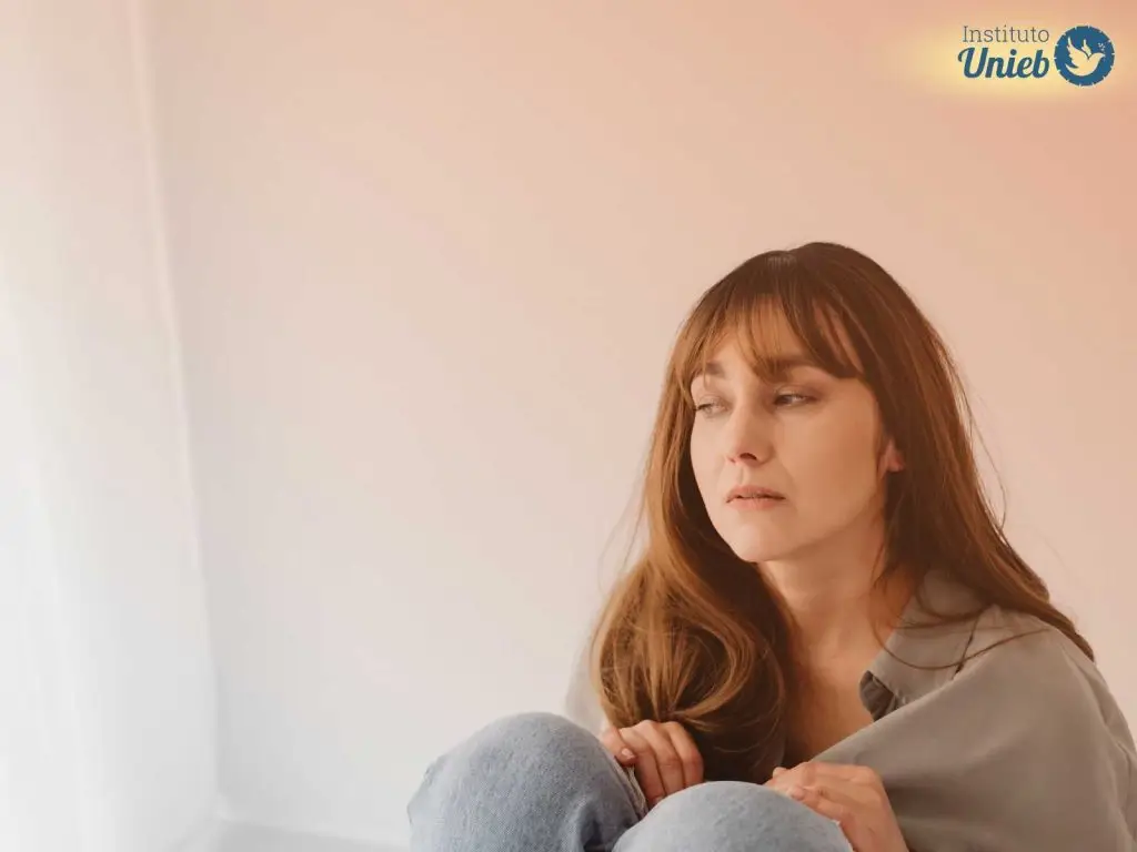 Mulher com cabelo solto vestindo roupa cinza e jeans sentada com mãos e corpo próximo ao joelho e expressão de tristeza