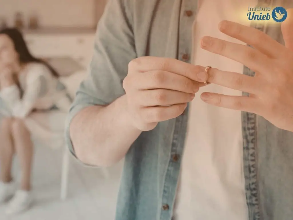 Homem vestindo camisa jeans azul claro e camiseta branca tirando aliança do dedo e ao fundo uma mulher lamentando