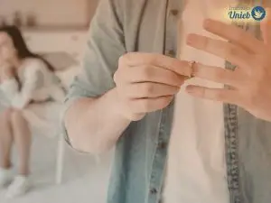 Homem vestindo camisa jeans azul claro e camiseta branca tirando aliança do dedo e ao fundo uma mulher lamentando