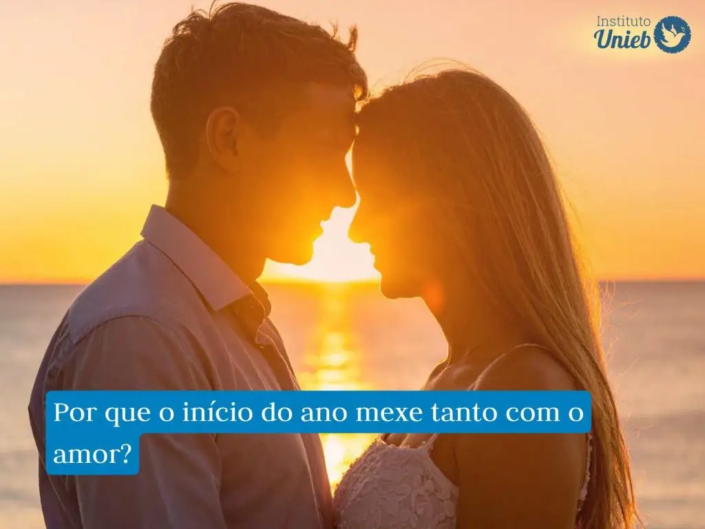 Homem e mulher de frente um para o outro encostando a cabeça um no outro em uma praia com pôr do sol ao fundo