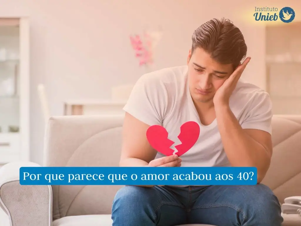 Homem sentado em sofá com cabeça apoiada em uma mão e com coração de papel partido ao meio na outra mão