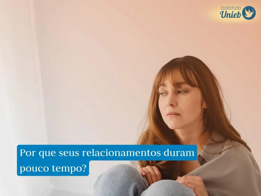 Mulher com cabelo solto vestindo roupa cinza e jeans sentada com mãos e corpo próximo ao joelho e expressão de tristeza