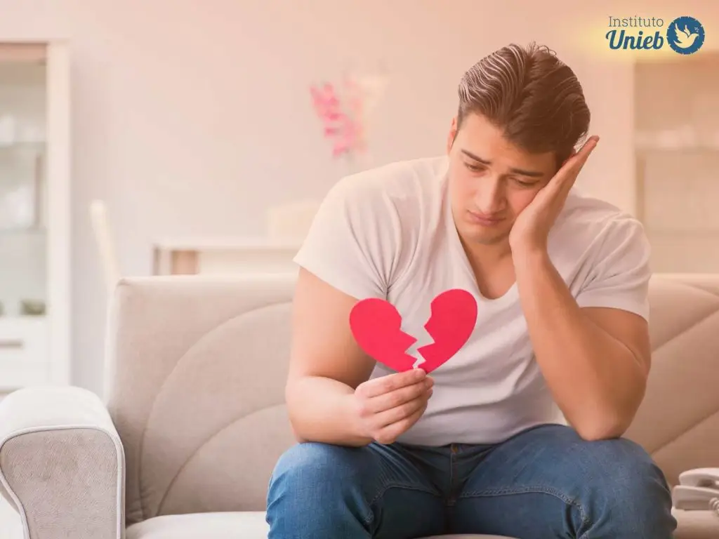 Homem sentado em sofá com cabeça apoiada em uma mão e com coração de papel partido ao meio na outra mão