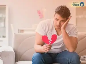 Homem sentado em sofá com cabeça apoiada em uma mão e com coração de papel partido ao meio na outra mão