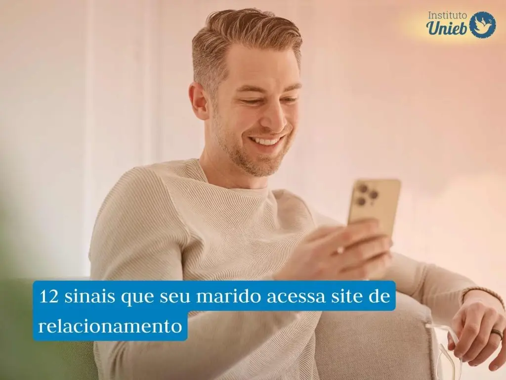 Homem vestindo blusa de manga longa de cor clara sentado em poltrona enquanto usa o celular e sorri, representando um marido que usa o celular escondido para acessar sites de relacionamento ou cometer traição virtual