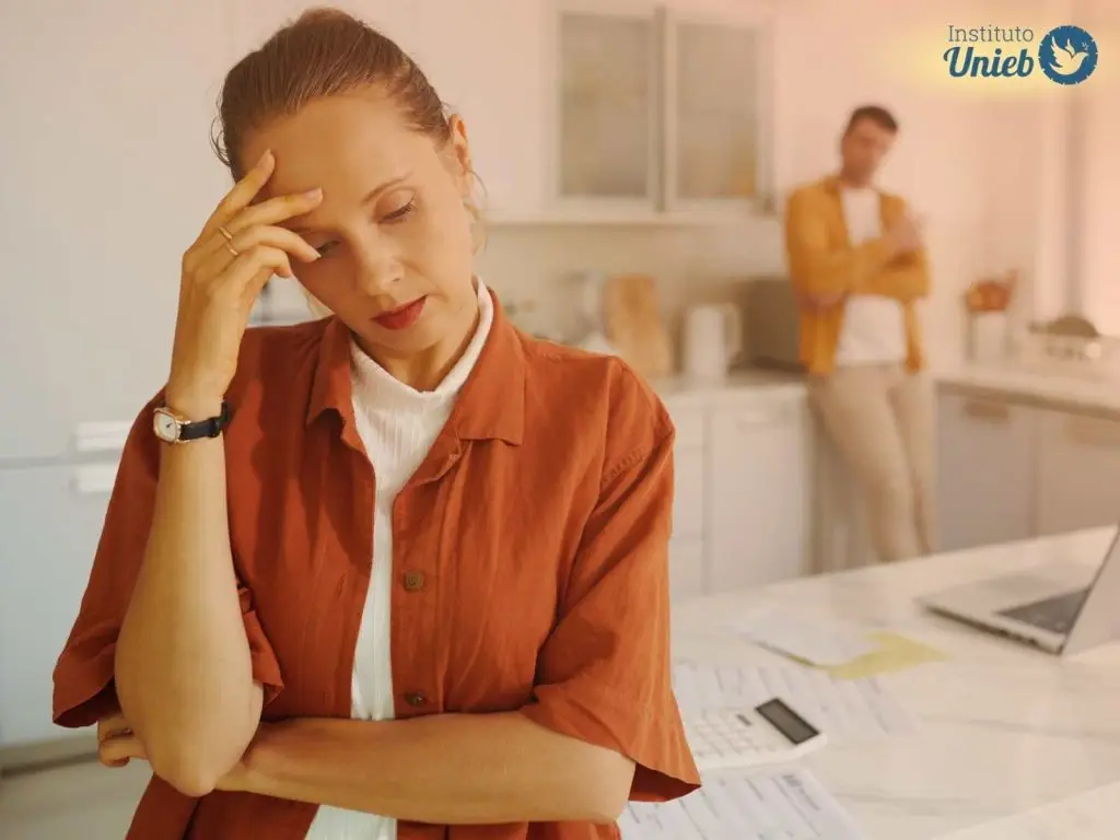 Mulher na cozinha com mão no rosto e expressão de frustração com homem ao fundo simbolizando o distanciamento emocional e a solidão no casamento