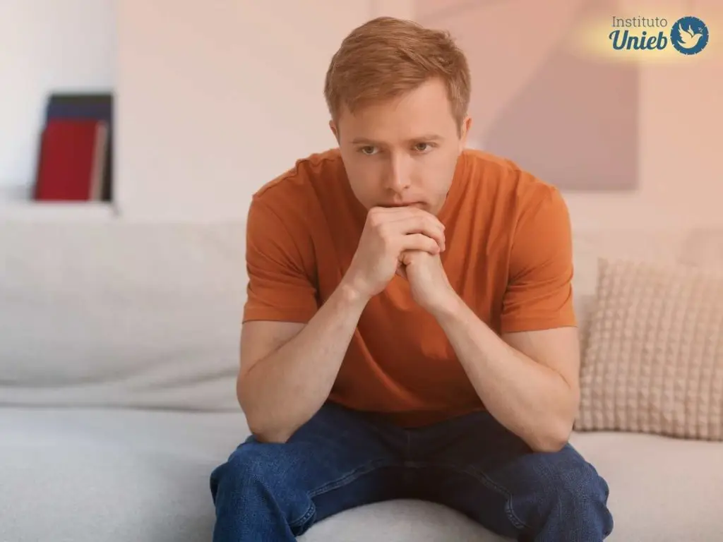 Homem vestindo camiseta laranja sentado no sofá com mãos fechadas e queixo apoiado indicando reflexão profunda como um homem obsessivo no amor