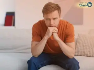 Homem vestindo camiseta laranja sentado no sofá com mãos fechadas e queixo apoiado indicando reflexão profunda como um homem obsessivo no amor