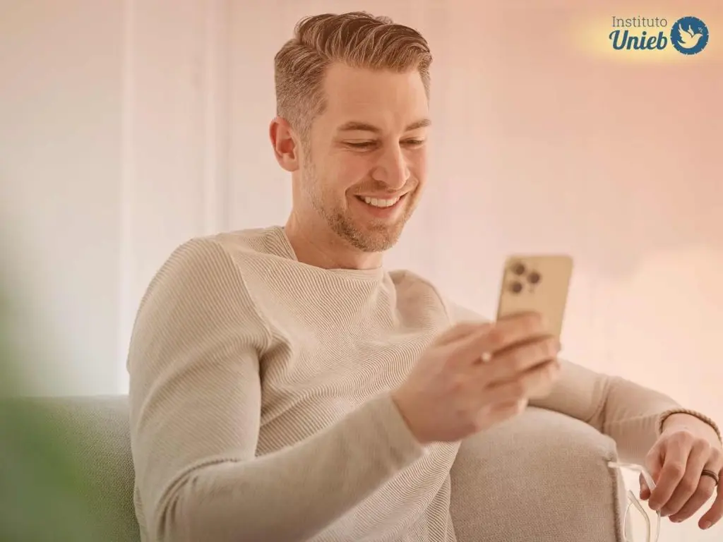 Homem vestindo blusa de manga longa de cor clara sentado em poltrona enquanto usa o celular e sorri, representando um marido que usa o celular escondido para acessar sites de relacionamento ou cometer traição virtual