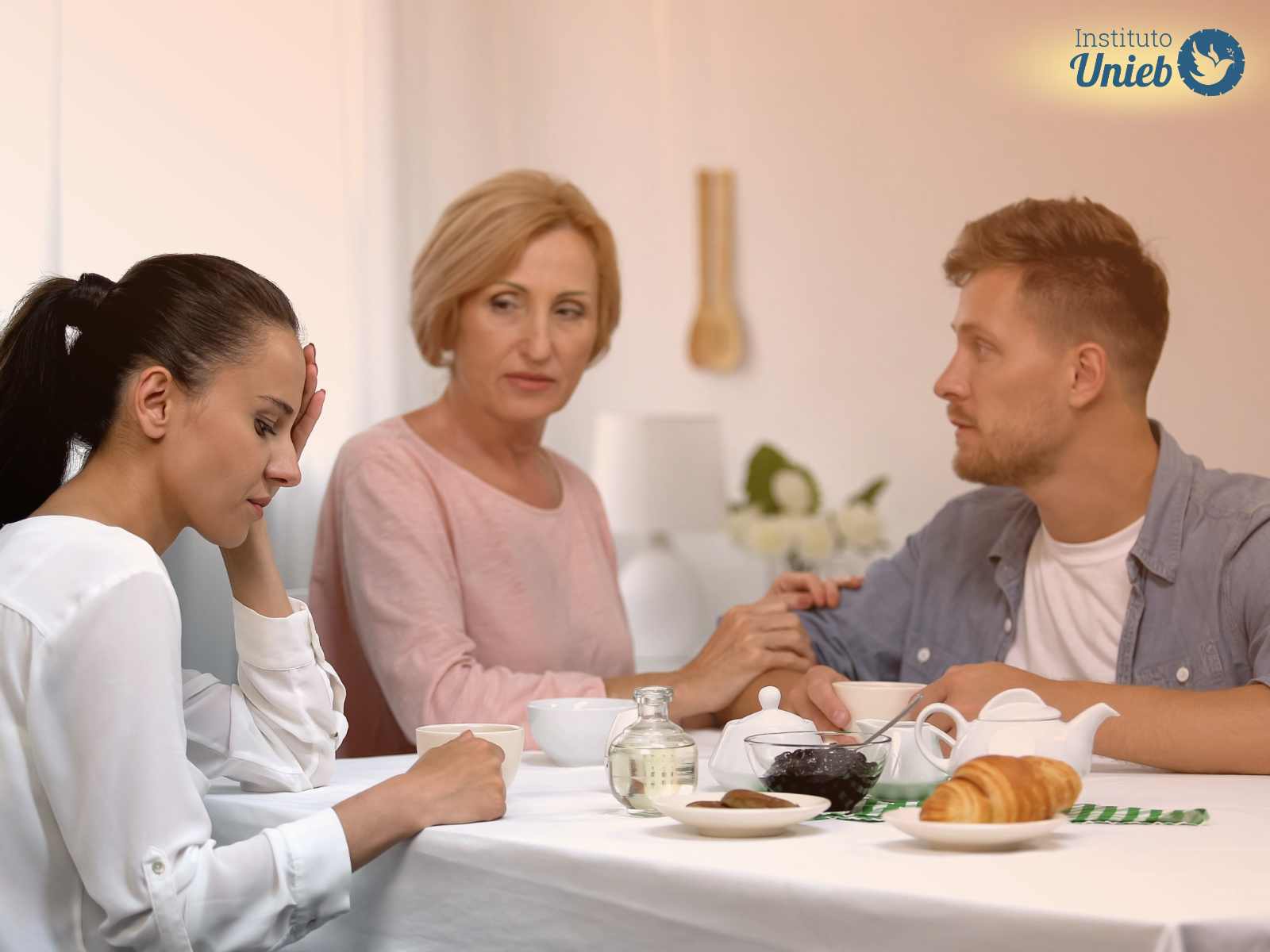Mulher com mão no rosto e expressão de frustração enquanto outra mulher mais velha, mãe do seu marido, consola o homem na mesa