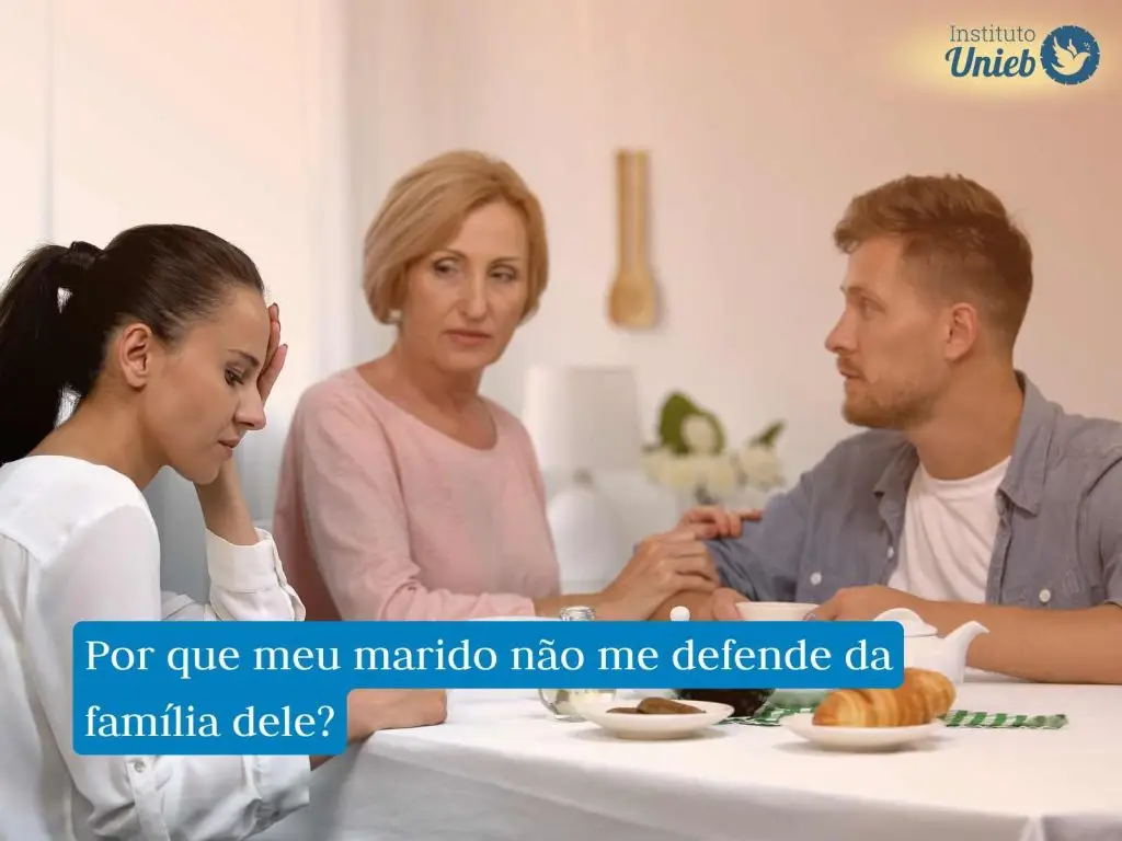 Mulher com mão no rosto e expressão de frustração enquanto outra mulher mais velha, mãe do seu marido, consola o homem na mesa