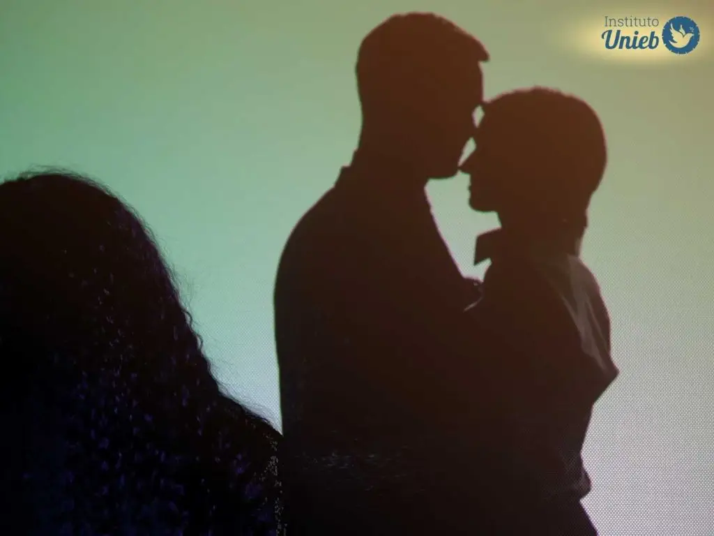 Casal beijando ao fundo enquanto mulher observa à frente indicando que seu parceiro está cometendo uma traição ao beijar outra mulher